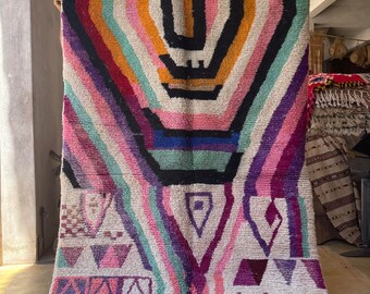 May include: A colorful Moroccan rug with a geometric pattern in shades of pink, purple, green, orange, and black. The rug has a white background and a fringe border.