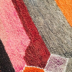 May include: A close-up of a colorful bath mat with a geometric pattern. The mat is made of a soft, plush material and features shades of pink, red, orange, gray, and purple.