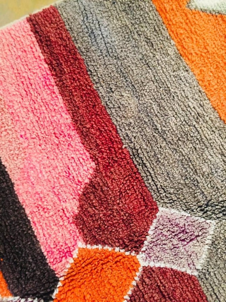 May include: A close-up of a colorful bath mat with a geometric pattern. The mat is made of a soft, plush material and features shades of pink, red, orange, gray, and purple.