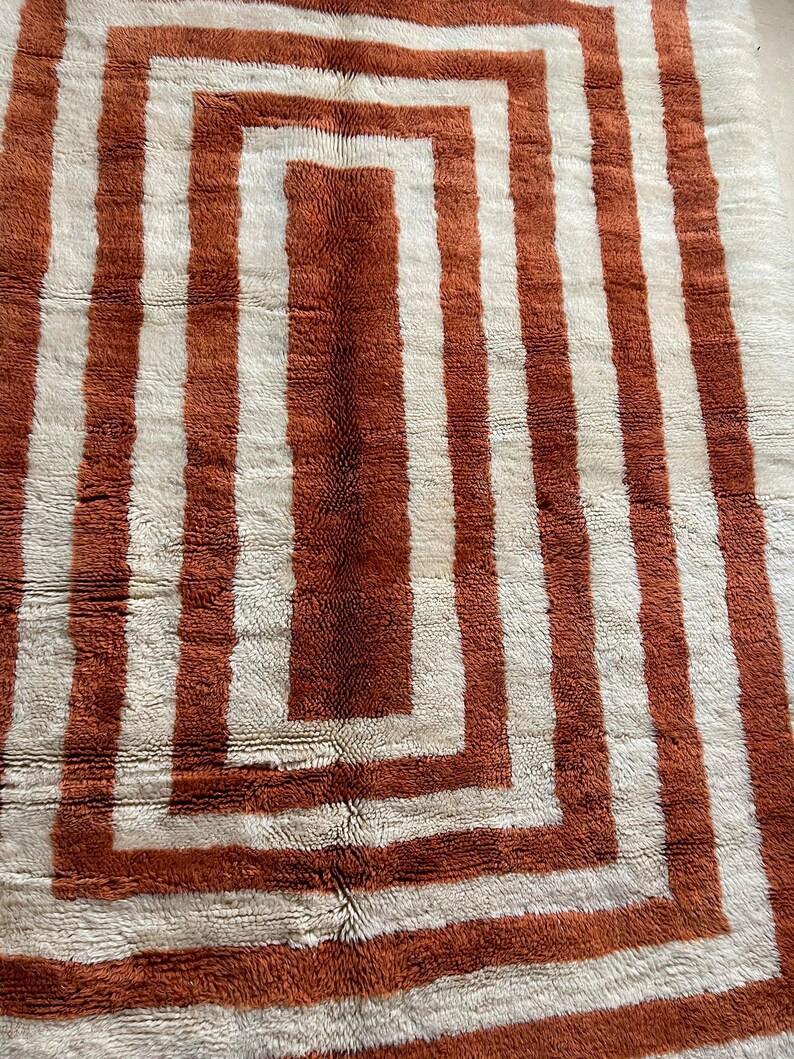 May include: A white and brown rug with a geometric pattern of concentric squares. The rug has a shaggy texture.
