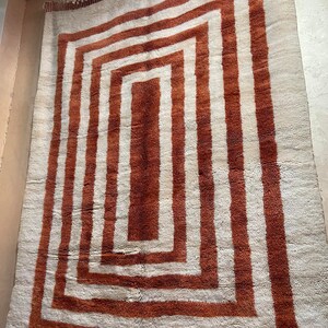 May include: A white rug with a large, concentric square pattern in shades of brown. The rug has a fringe border.