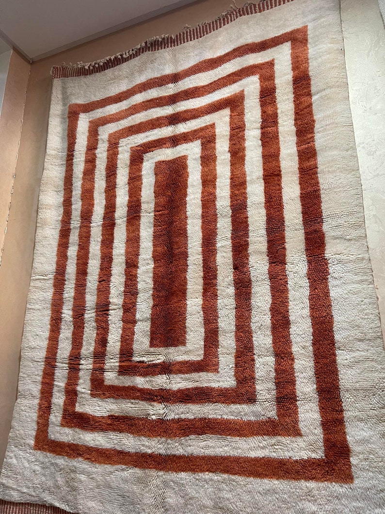 May include: A white rug with a large, concentric square pattern in shades of brown. The rug has a fringe border.
