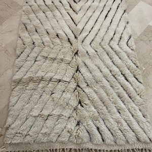 K&ouml;nnte beinhalten: Ein wei&szlig;er und brauner marokkanischer Teppich mit geometrischem Muster. Der Teppich hat einen Fransenrand und ist aus Wolle.