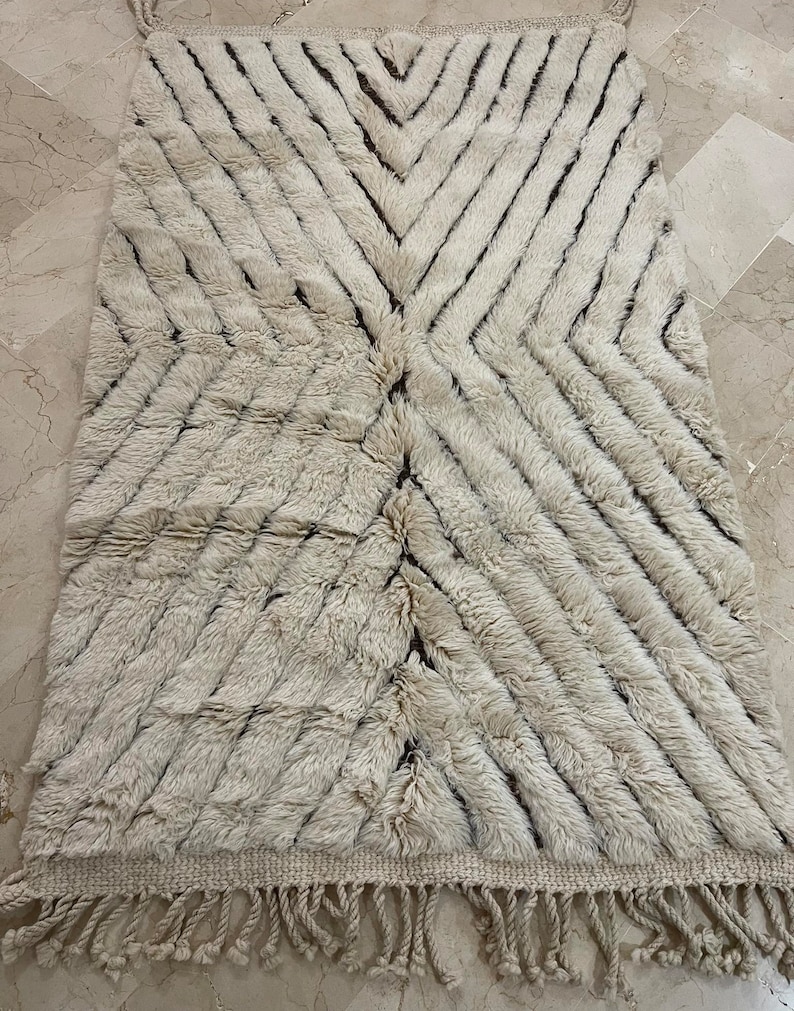 K&ouml;nnte beinhalten: Ein wei&szlig;er und brauner marokkanischer Teppich mit geometrischem Muster. Der Teppich hat einen Fransenrand und ist aus Wolle.