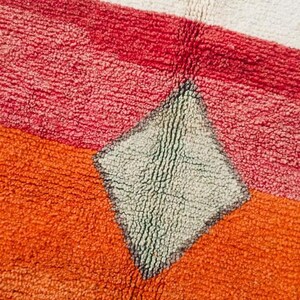 May include: Close-up of a colorful rug with a diamond-shaped pattern in the center. The rug is made of a soft, textured material and features shades of red, orange, and white.
