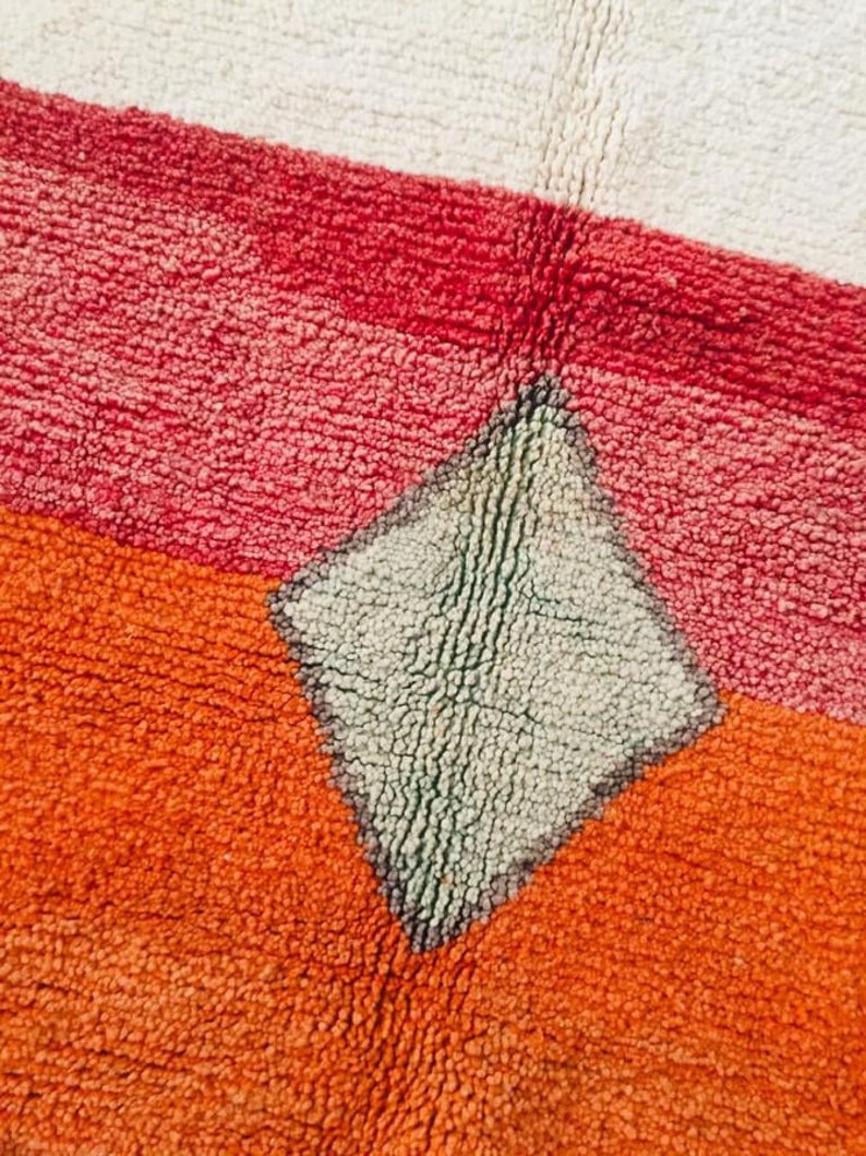 May include: Close-up of a colorful rug with a diamond-shaped pattern in the center. The rug is made of a soft, textured material and features shades of red, orange, and white.