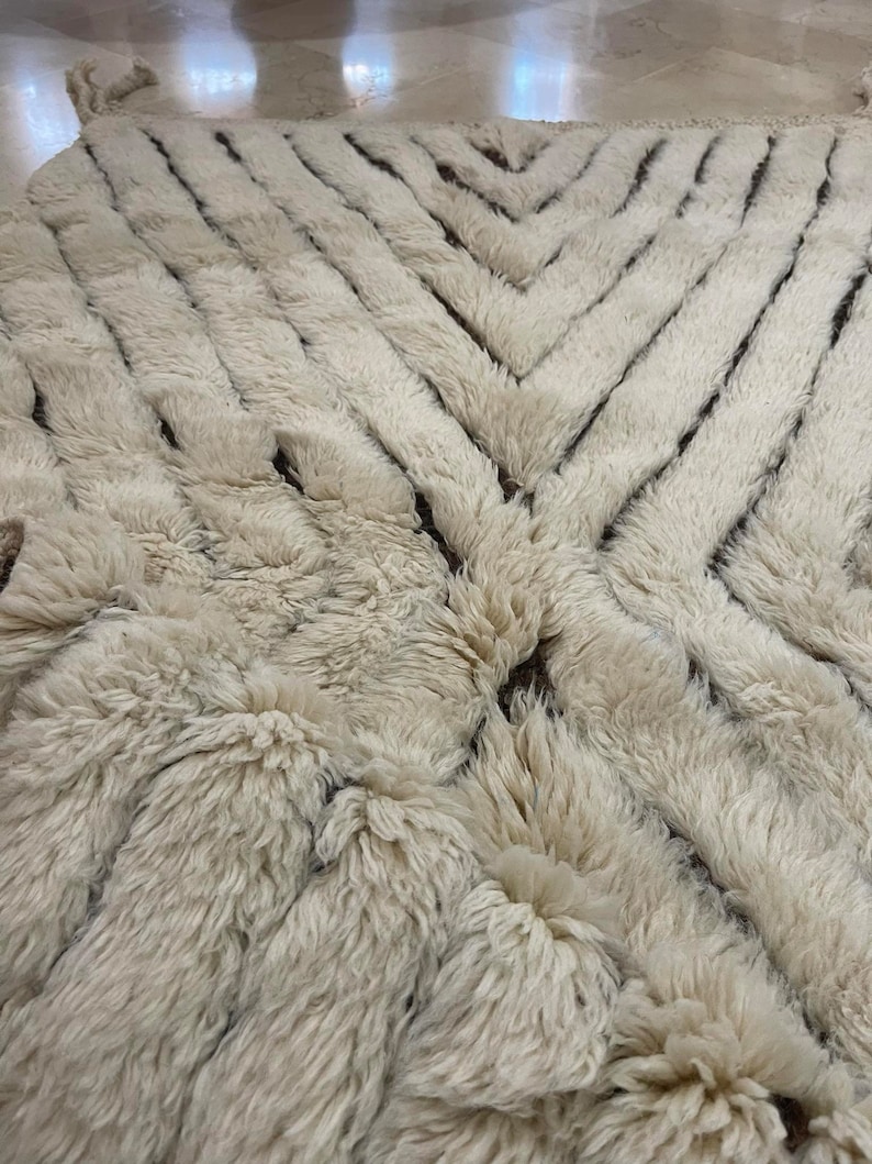 K&ouml;nnte beinhalten: Ein wei&szlig;er Shaggy-Teppich mit einem geometrischen Muster aus braunen Linien. Der Teppich hat eine weiche, flauschige Textur.