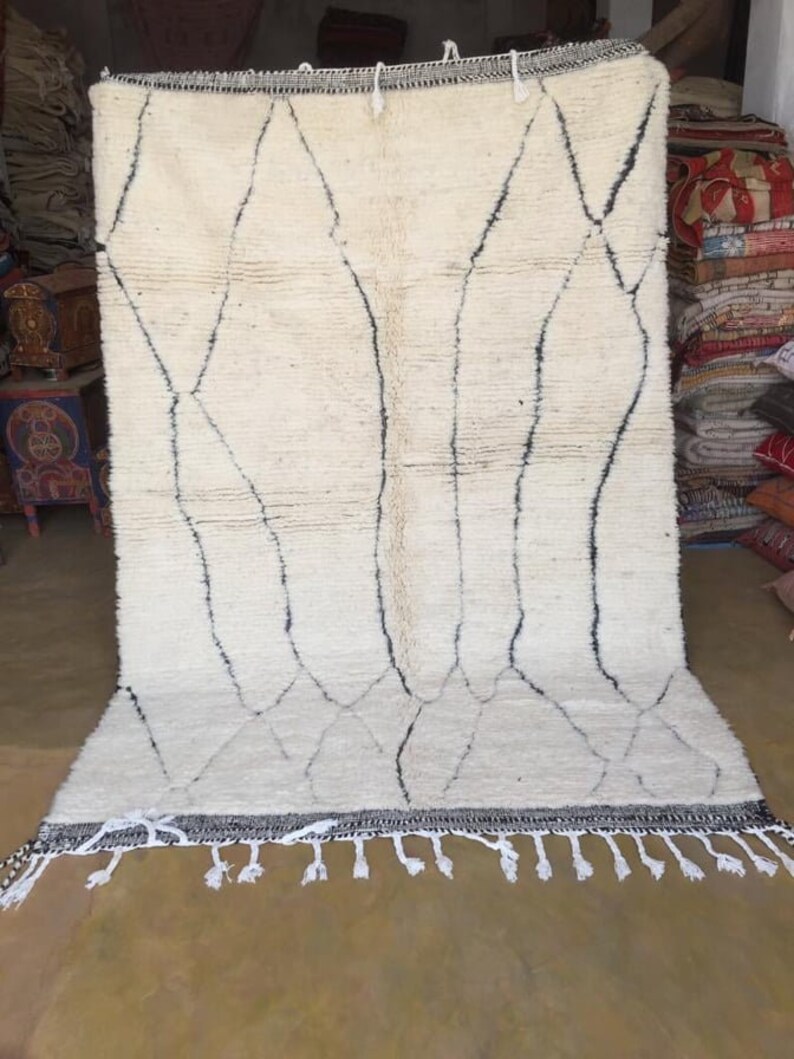 May include: A white Moroccan rug with a black diamond pattern and fringe. The rug is made of wool and is hand-woven.