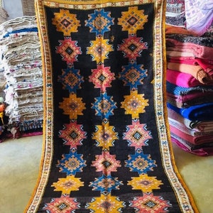 May include: A black Moroccan rug with a geometric pattern in shades of yellow, blue, pink, and white. The rug has a fringed border and is folded in half.