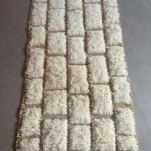 May include: A white and beige rectangular rug with a geometric pattern of squares. The rug is made of a fluffy, textured material.
