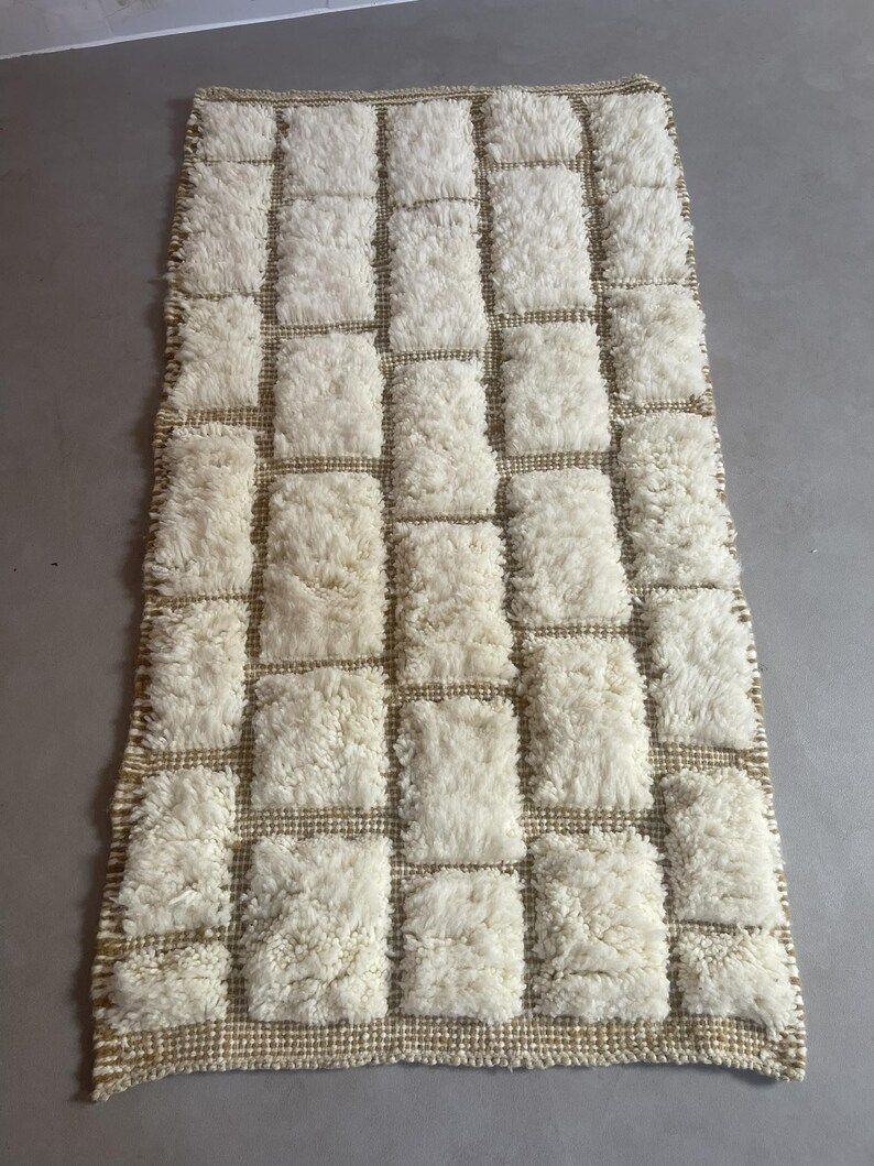 May include: A white and beige rectangular rug with a geometric pattern of squares. The rug is made of a fluffy, textured material.