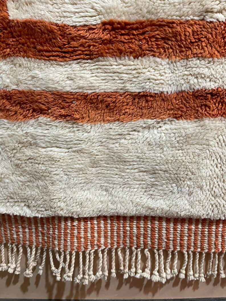 May include: A white and orange striped rug with a fringe border. The rug is made of a soft, fluffy material.
