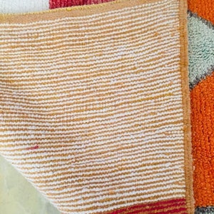 May include: The back side of a colorful rug with a white, red, orange, and green geometric pattern. The rug has a tan and white striped backing.