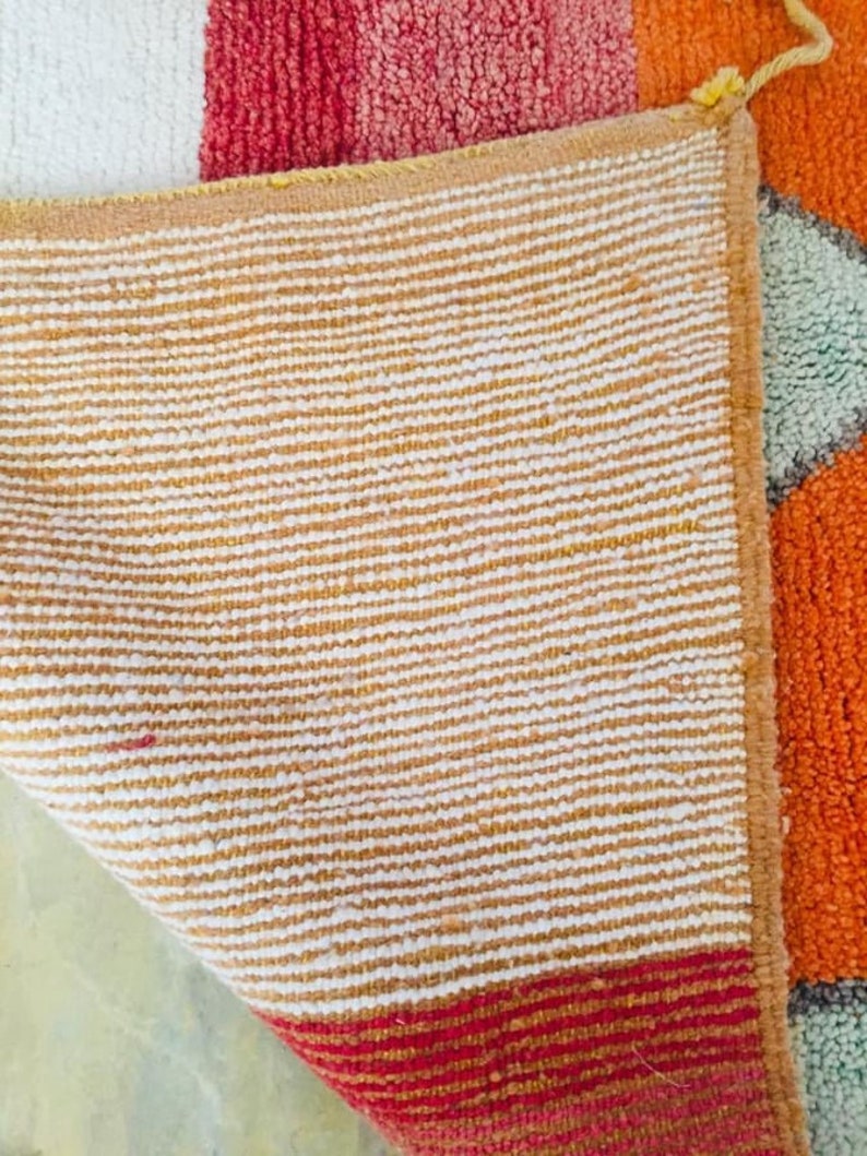 May include: The back side of a colorful rug with a white, red, orange, and green geometric pattern. The rug has a tan and white striped backing.