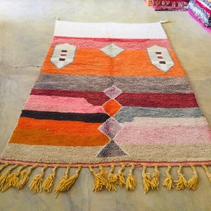 May include: A colorful Moroccan rug with a geometric pattern in shades of orange, pink, gray, and white. The rug has a fringe border and is made of wool.