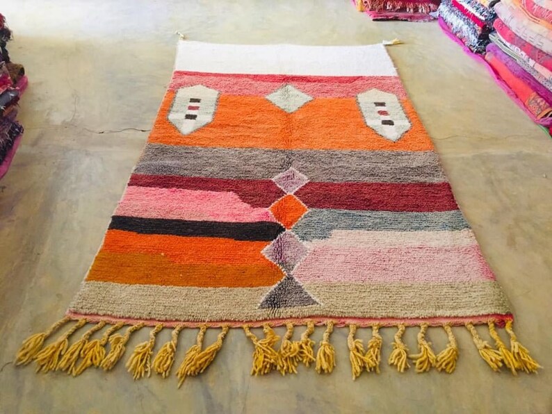 May include: A colorful Moroccan rug with a geometric pattern in shades of orange, pink, gray, and white. The rug has a fringe border and is made of wool.