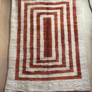 May include: A white rug with a geometric pattern of concentric brown squares. The rug has a fringe border.