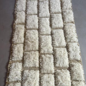 May include: A white and beige rectangular rug with a geometric pattern of squares. The rug is made of a fluffy, textured material.