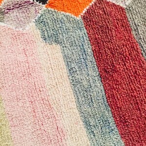 May include: A close-up of a colorful rug with a geometric pattern. The rug is made of a soft, fuzzy material and features shades of pink, orange, red, blue, and gray.