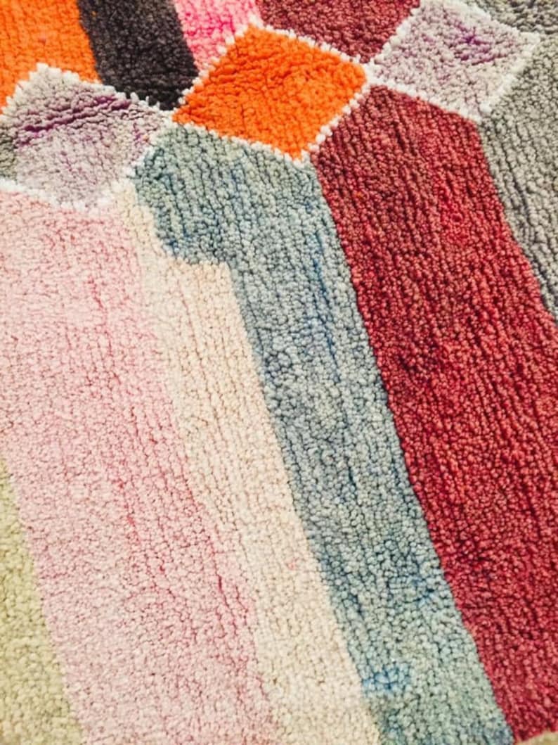 May include: A close-up of a colorful rug with a geometric pattern. The rug is made of a soft, fuzzy material and features shades of pink, orange, red, blue, and gray.