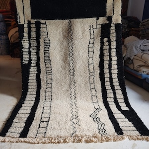May include: A white and black Moroccan rug with a geometric pattern. The rug has a fringe border and is made of wool.