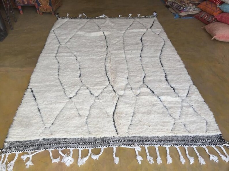 May include: White Moroccan rug with a geometric pattern in black. The rug has a fringe border.