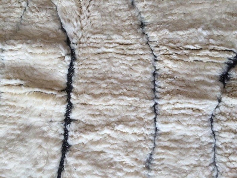 May include: Close-up of a white, fluffy, woven textile with black stitching. The texture is soft and plush, resembling a sheepskin rug.