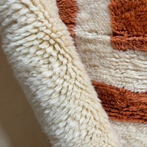 May include: Close-up of a white and orange striped rug with a fluffy texture. The rug is made of wool and has a hand-woven look.