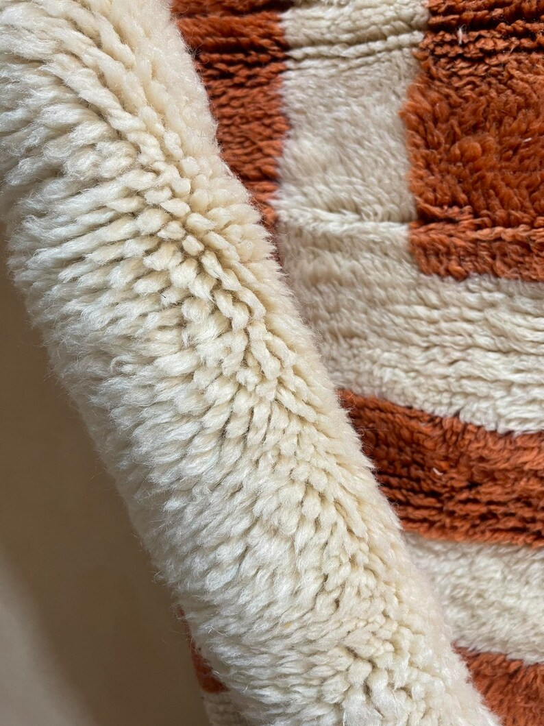 May include: Close-up of a white and orange striped rug with a fluffy texture. The rug is made of wool and has a hand-woven look.