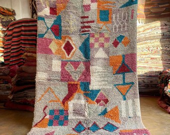 May include: A colorful Moroccan rug with geometric patterns in shades of red, orange, blue, and pink. The rug has a white background and is made of wool. It has a fringe border.