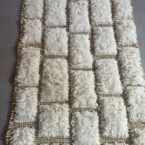 May include: White and beige rectangular rug with a tufted texture. The rug has a grid pattern with squares of white tufting and beige woven lines.