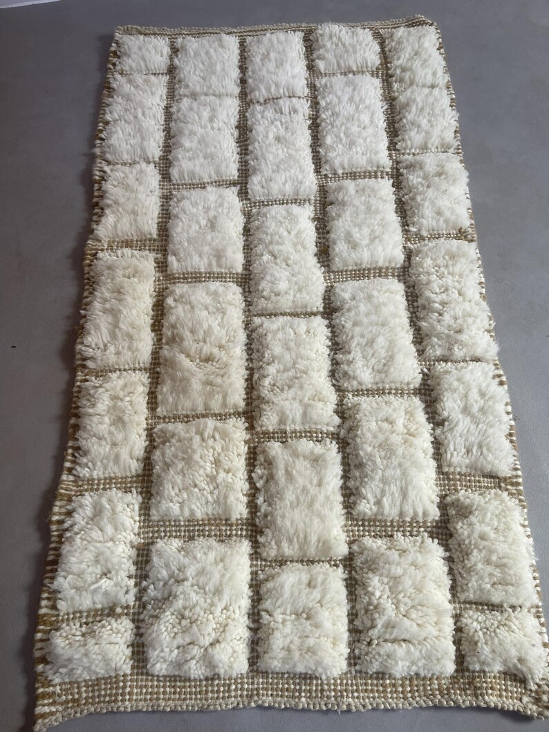 May include: A white and beige rectangular rug with a geometric pattern of squares. The rug is made of a fluffy, textured material.
