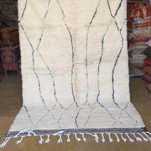 May include: A white Moroccan rug with a black diamond pattern and fringe. The rug is hanging on a wall in a shop.