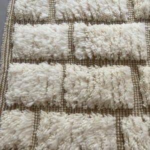 May include: Close-up of a white and brown woven rug with a textured, fluffy surface. The rug has a geometric pattern of squares.
