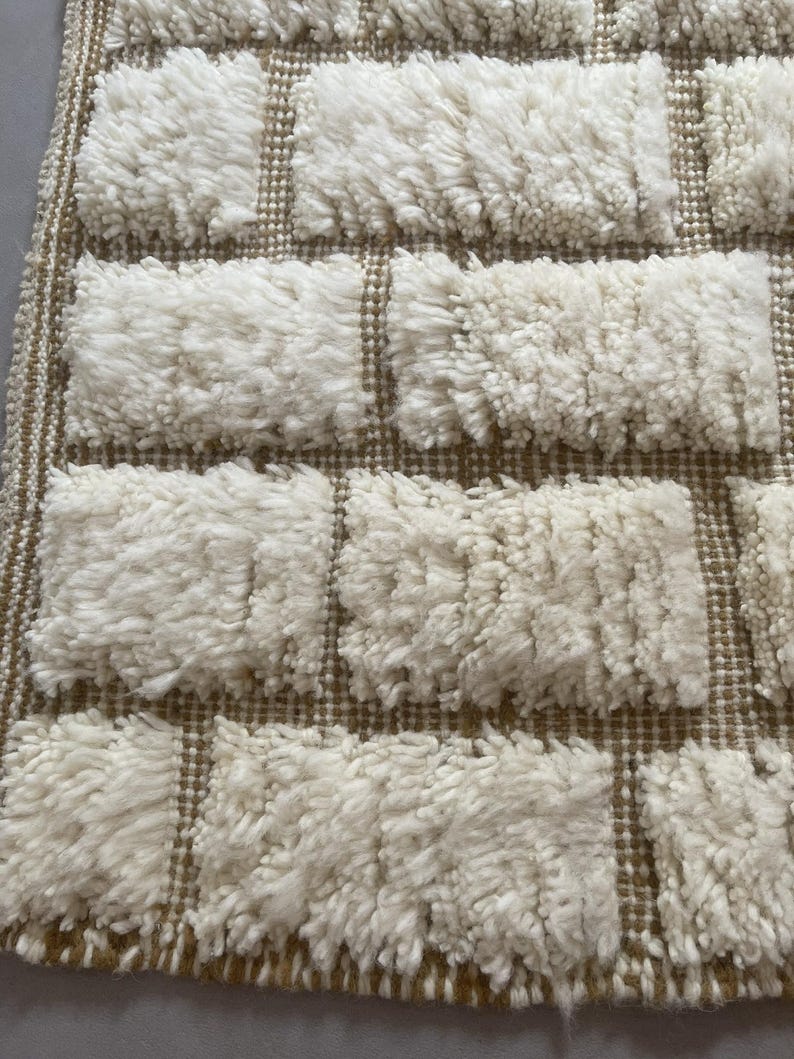 May include: Close-up of a white and brown woven rug with a textured, fluffy surface. The rug has a geometric pattern of squares.