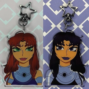 May include: Two clear acrylic keychains featuring cartoon portraits of female characters. The keychain on the left features a red-haired character with green eyes and a blue and purple outfit. The keychain on the right features a black-haired character with purple eyes and a blue and purple outfit.