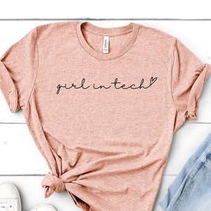 May include: A light pink t-shirt with the text "girl in tech" in black lettering with a small heart symbol. The shirt is laid flat on a white wooden surface with a pair of white sneakers and a pair of light blue jeans.