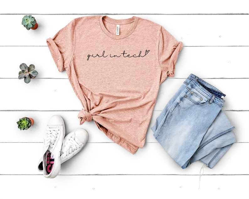 May include: A light pink t-shirt with the text "girl in tech" in black lettering with a small heart symbol. The shirt is laid flat on a white wooden surface with a pair of white sneakers and a pair of light blue jeans.