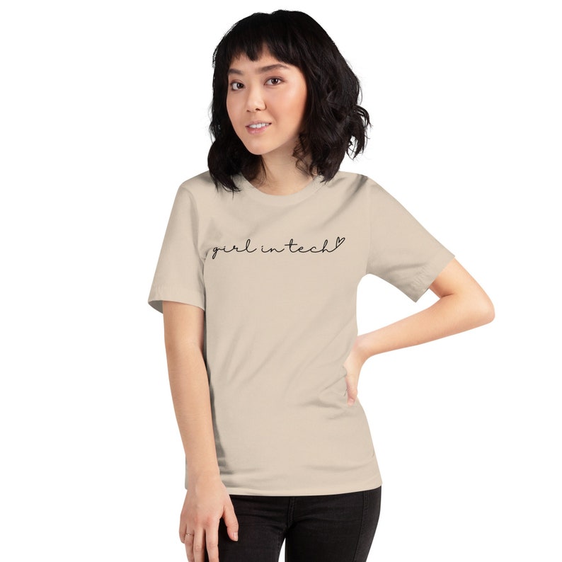 May include: A beige t-shirt with the text "girl in tech" written in black cursive with a small heart symbol after the word "tech".