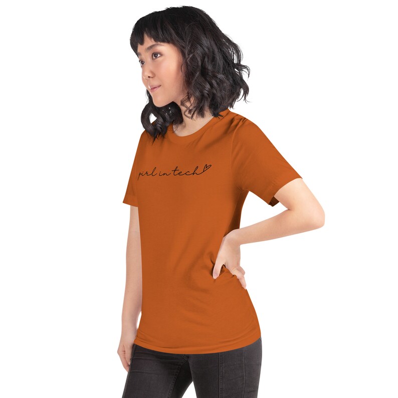 May include: A woman wearing a rust-colored t-shirt with the text "girl in tech" and a small heart symbol.