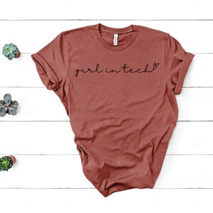 May include: A rust-colored t-shirt with the text "girl in tech" in black lettering with a small heart symbol.
