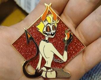 May include: Gold-colored enamel pin with a red glitter background. The pin features a cartoon character with yellow hair, horns, and a tail. The character is wearing a black choker and fishnet stockings. The character is holding a red apple in their hand.