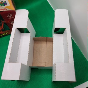 May include: A white cardboard insert for a Nintendo 64 game box. The insert has a rectangular base with two vertical supports and a central compartment. The background is a green surface.