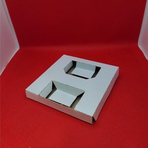 May include: White cardboard box with two cut-out rectangular holes. The box is folded and has a slight crease in the middle.