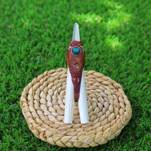 May include: A white crystal point wrapped in brown leather with turquoise and red gemstone accents. The crystal is sitting on a woven straw mat.