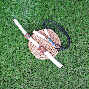 May include: Two white bone pipes with decorative metal accents and turquoise stones. The pipes are on a woven straw mat with a black cord necklace.