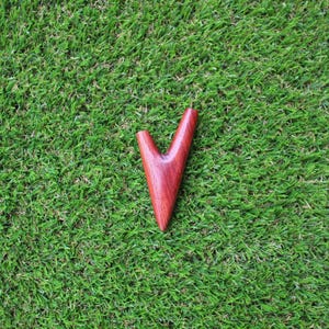 May include: A wooden V-shaped pipe, made of reddish-brown wood, is lying on a green grassy surface.