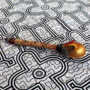 May include: A wooden rattle with a gold-painted gourd, green gemstone accents, and feathers. The rattle is on a white fabric with a black geometric pattern.