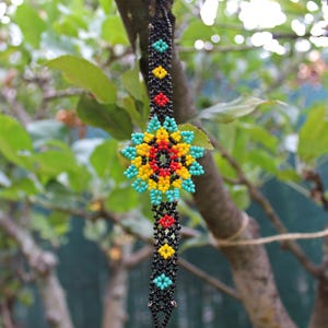 May include: A beaded bracelet with a starburst design. The bracelet features a central starburst in turquoise, yellow, red, and black beads. The band is black with turquoise, yellow, and red accents. The bracelet is hanging from a tree branch.