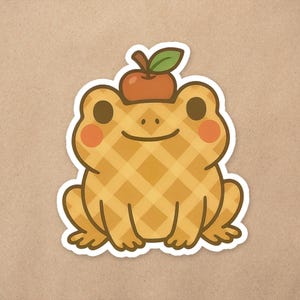 May include: A cheerful cartoon frog sticker with a waffle-like pattern in shades of yellow and brown. The frog has rosy cheeks and a small apple with a green leaf on its head. The sticker is outlined in white.